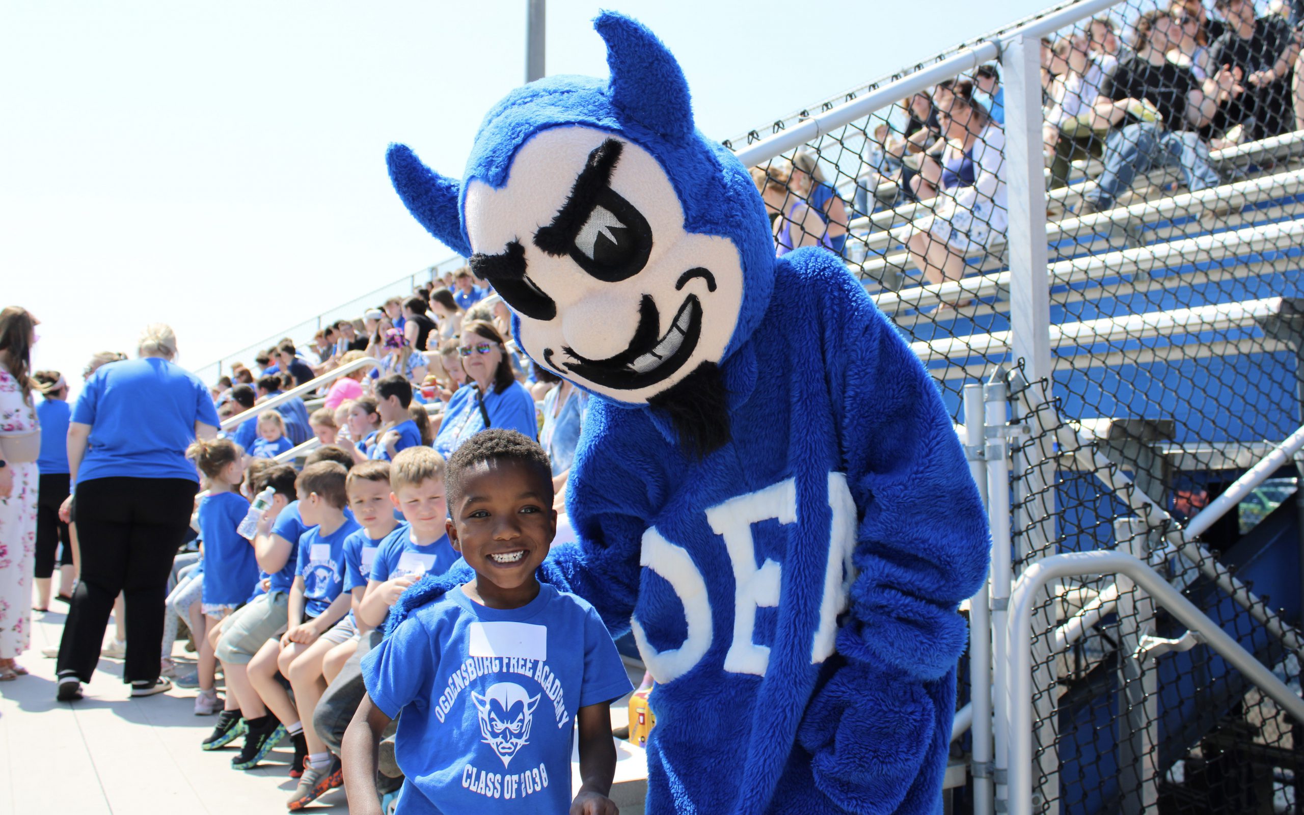 Young male student wearing a blue and white t-shirt smiling next to a blue and white devil mascot