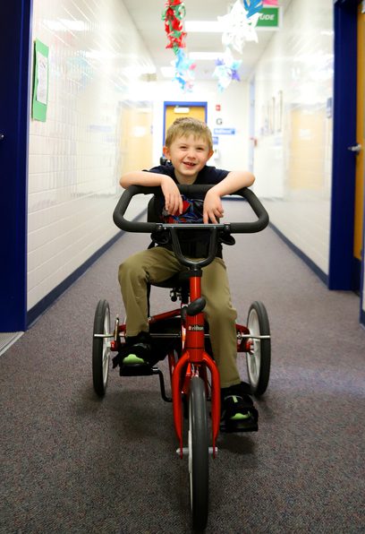 An elementary student sits on a tricycle bike in the school hallway.