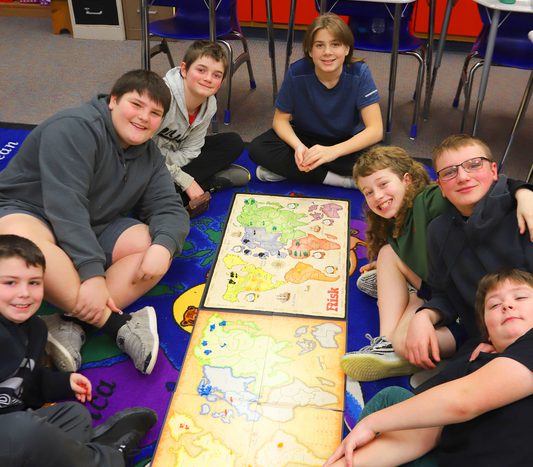 A group of students gather around a floor game of Risk.