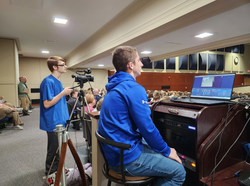 Two high school students film and monitor technology from the back of a packed auditorium.