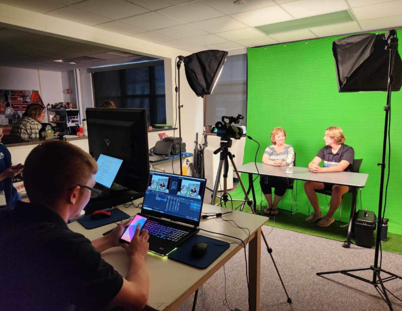 A student monitors equipment recording two people seated in front of a green screen.