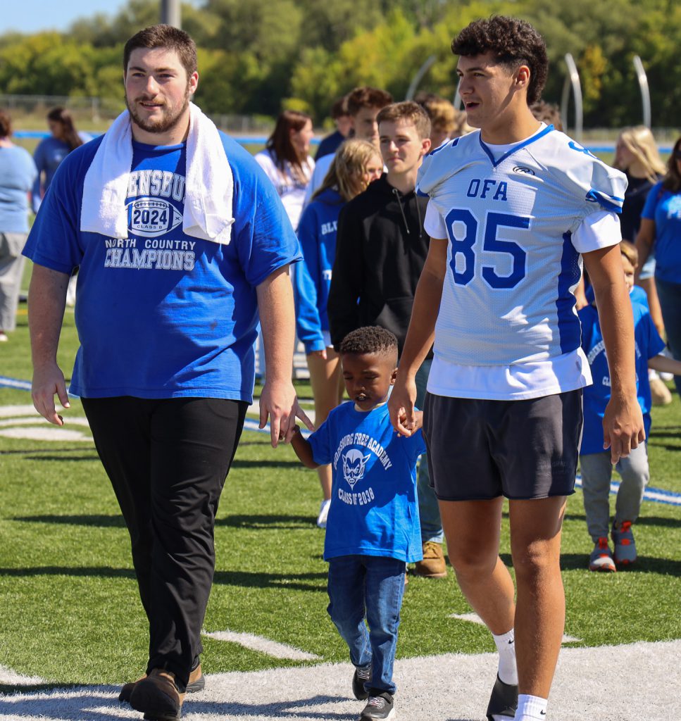 Two football players walk an elementary student across the playing field.