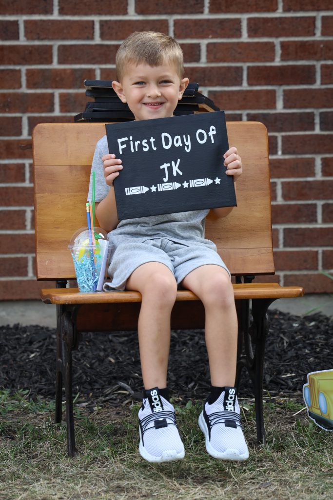 A young child seated and holding a sign that says "first day of JK".