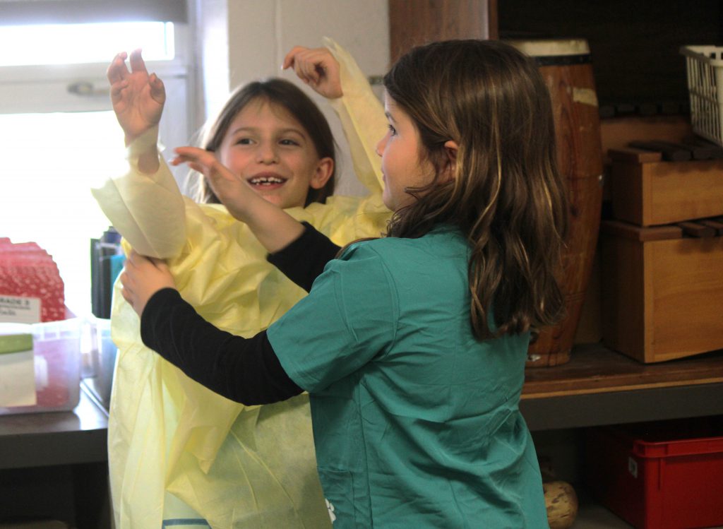 Female elementary student helping fellow female elementary student put on hospital scrubs