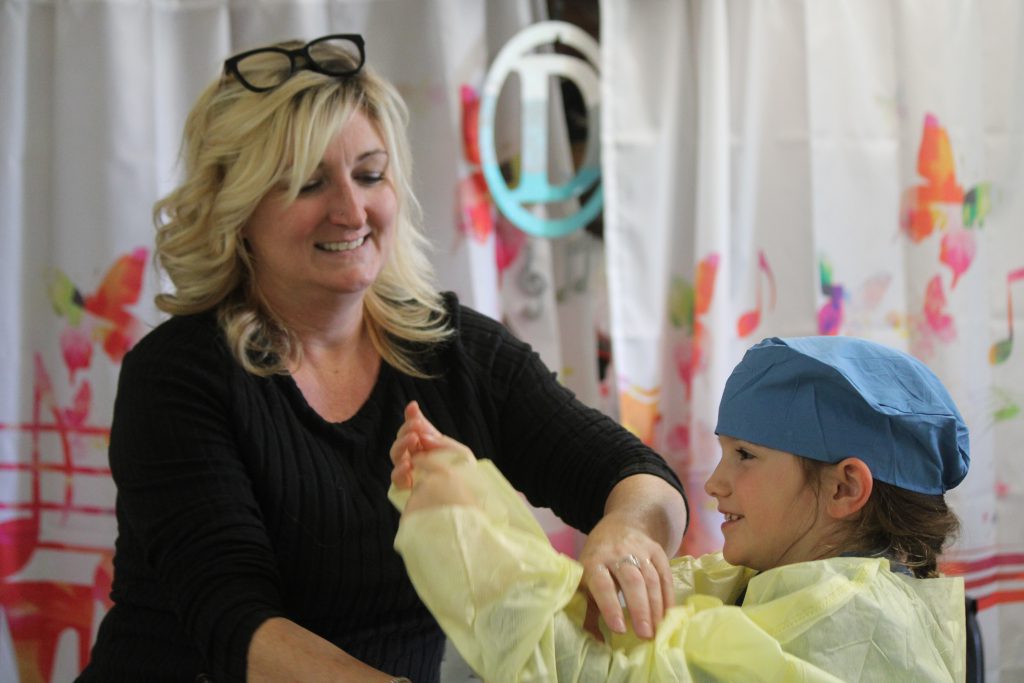 Female elementary teacher assisting female elementary student in putting on hospital scrubs