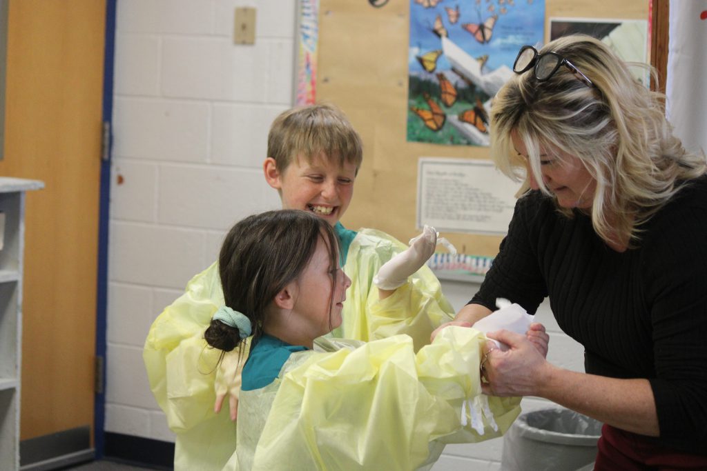 Female elementary teacher helping elementary students put on hospital scrubs