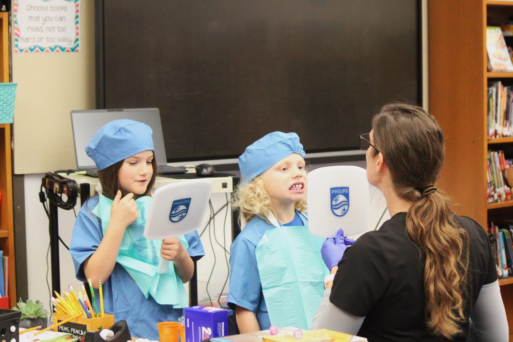 Male and female elementary students check out teeth in mirror while female dental staff member supervises