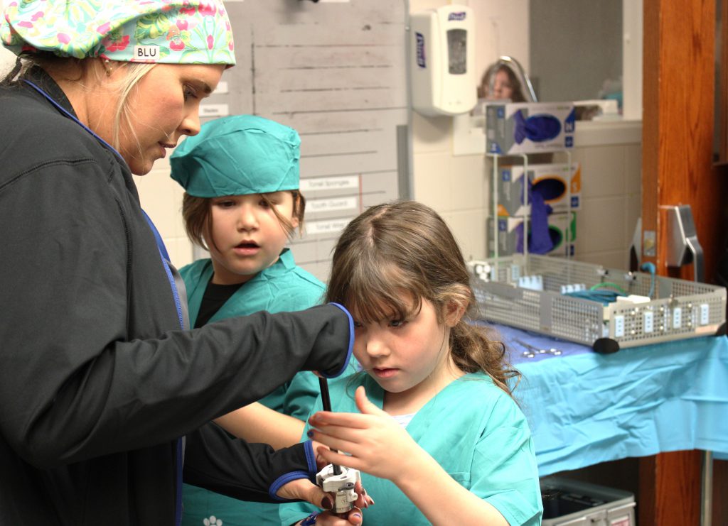 Elementary student touching medical device with nurse assistance with another student looking on