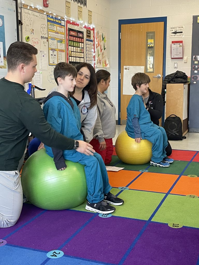 Elementary students wearing green hospital scrubs balancing on a ball with adults assisting
