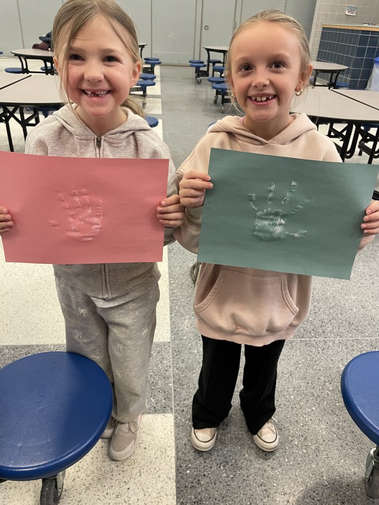 Two female elementary students smiling while holding up pink and green papers with their hand prints