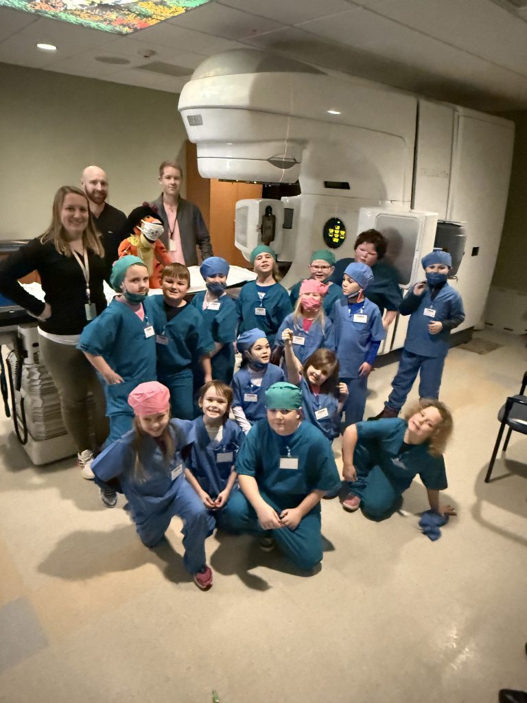 Small group of elementary students wearing blue hospital scrubs near hospital equipment with adults