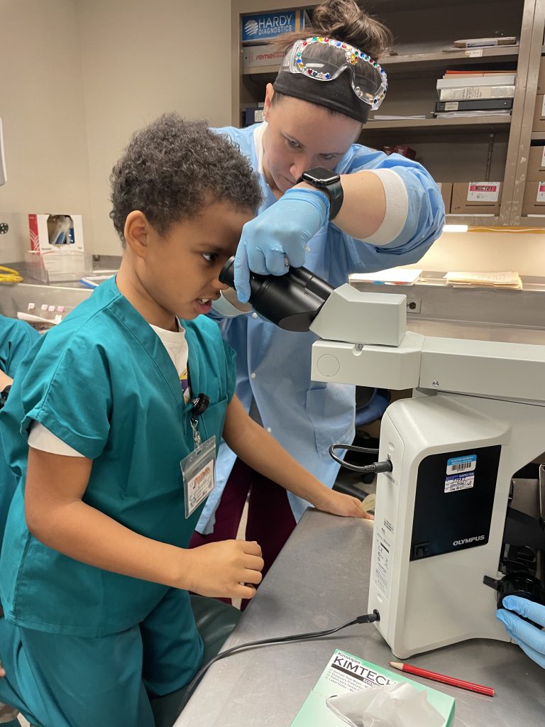 Male elementary student wearing green hospital scrubs looking through hospital equipment with female adult assisting