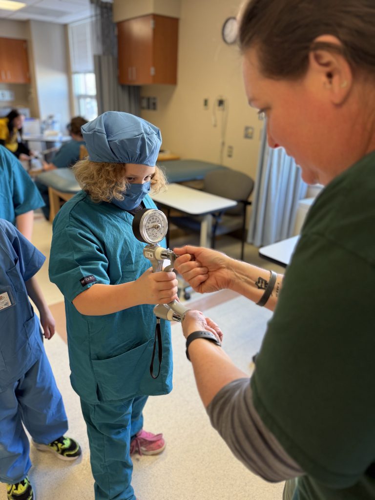 Elementary student wearing green hospital scrubs using hospital equipment with a female adult assisting