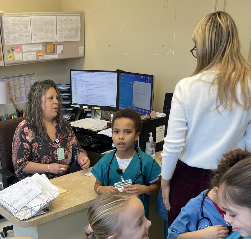 Male elementary student wearing green hospital scrubs with two adult females
