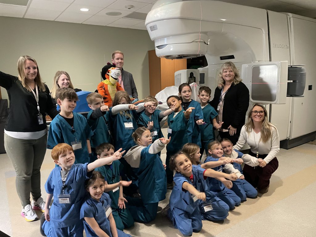 Small group of elementary students wearing blue and green hospital scrubs pointing next to hospital equipment with adults