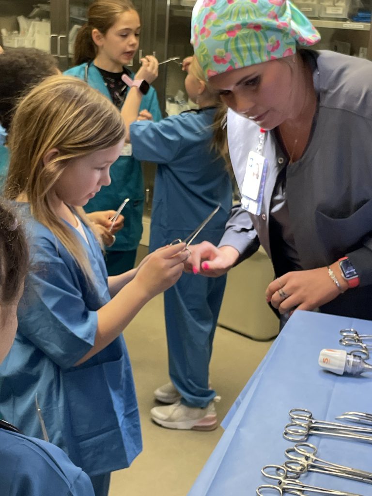 Female elementary student wearing blue hospital scrubs using medical equipment with adult female assisting