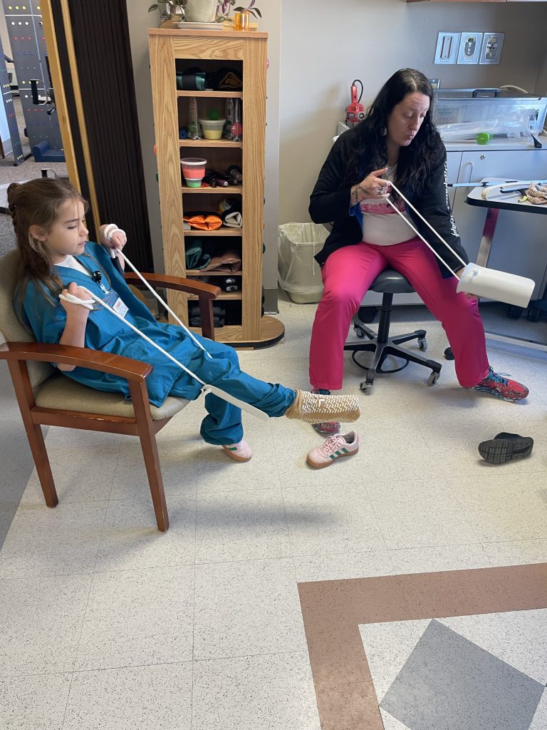 Female elementary student wearing green hospital scrubs using medical equipment with female adult assisting