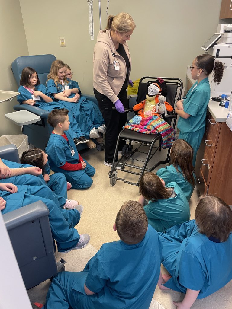 Small group of elementary students wearing blue and green hospital scrubs looking at plush tiger with adult female