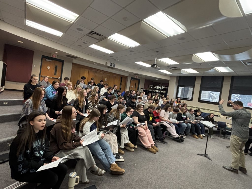 Large group of male and female students holding books with an adult male conducting in front of a classroom