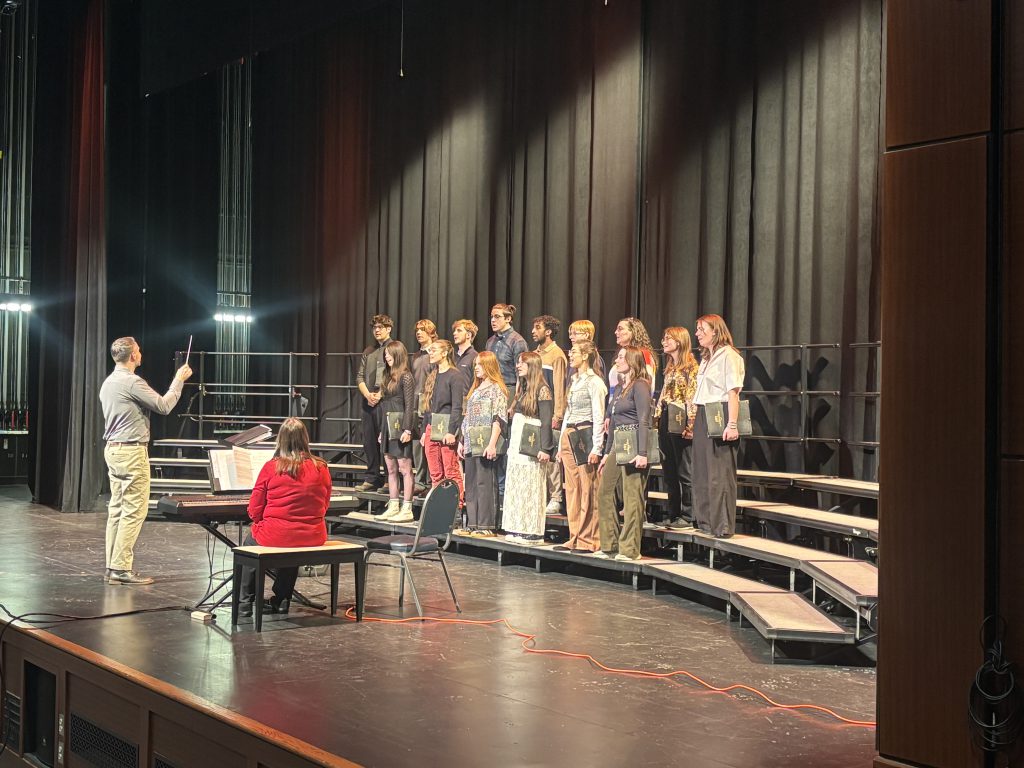 Small group of male and female students standing on risers performing with an adult male conducting in front of them