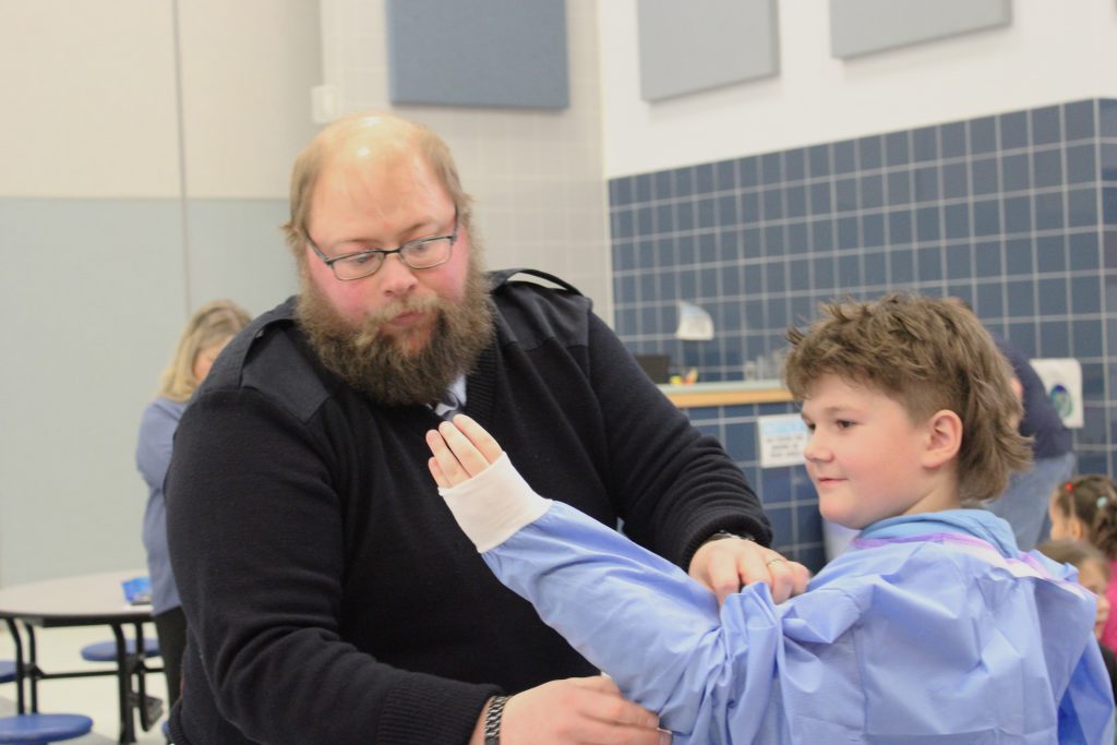 Adult male teacher helping male elementary student put on blue hospital scrubs
