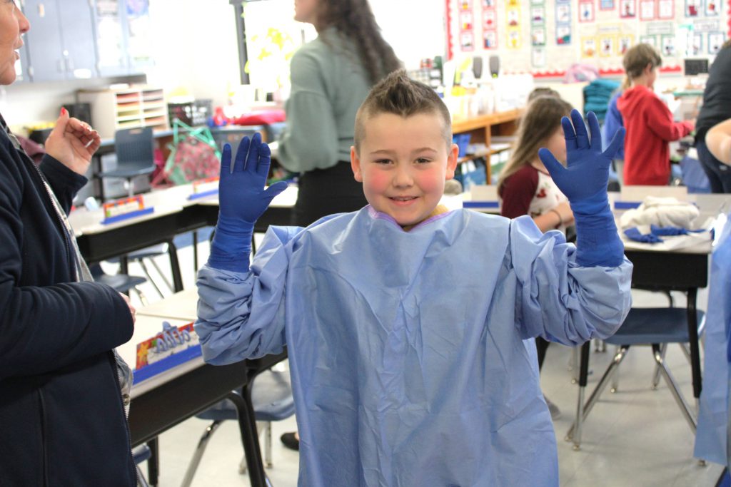 Male elementary student smiling wearing blue hospital scrubs and gloves