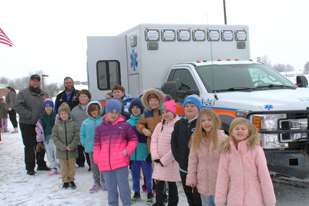 Small group of elementary students wearing jackets smiling by an ambulance with adults