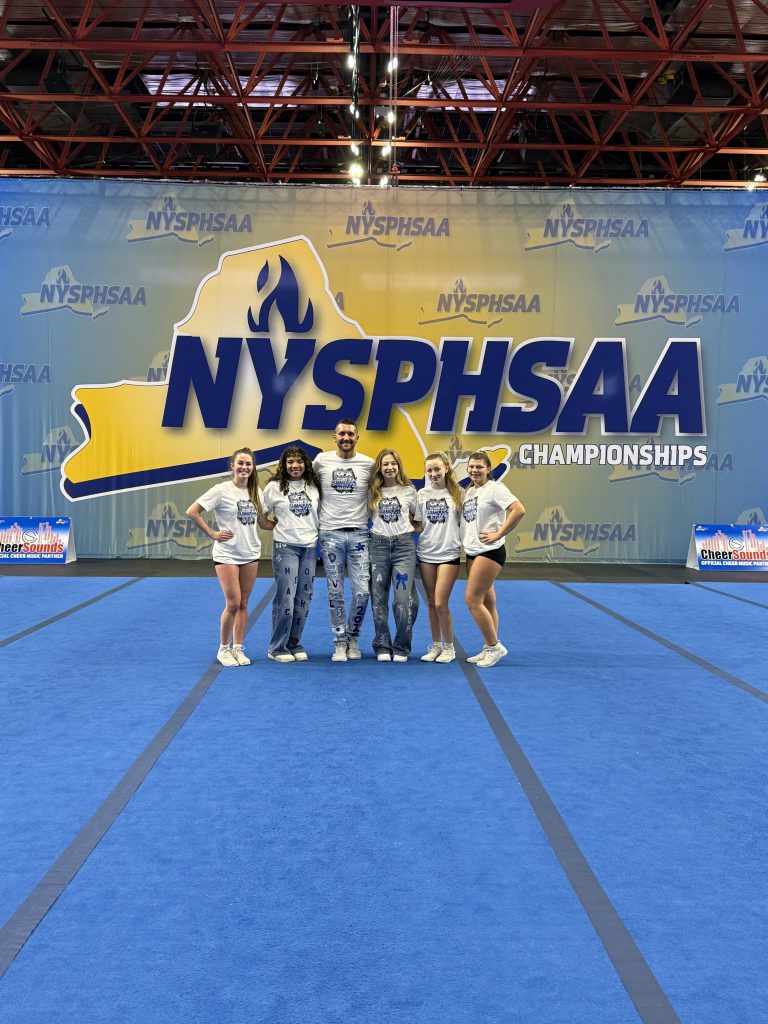 Small group of female high school cheerleaders with a female and male coach wearing white at the New York State Public High School Athletic Association state championships