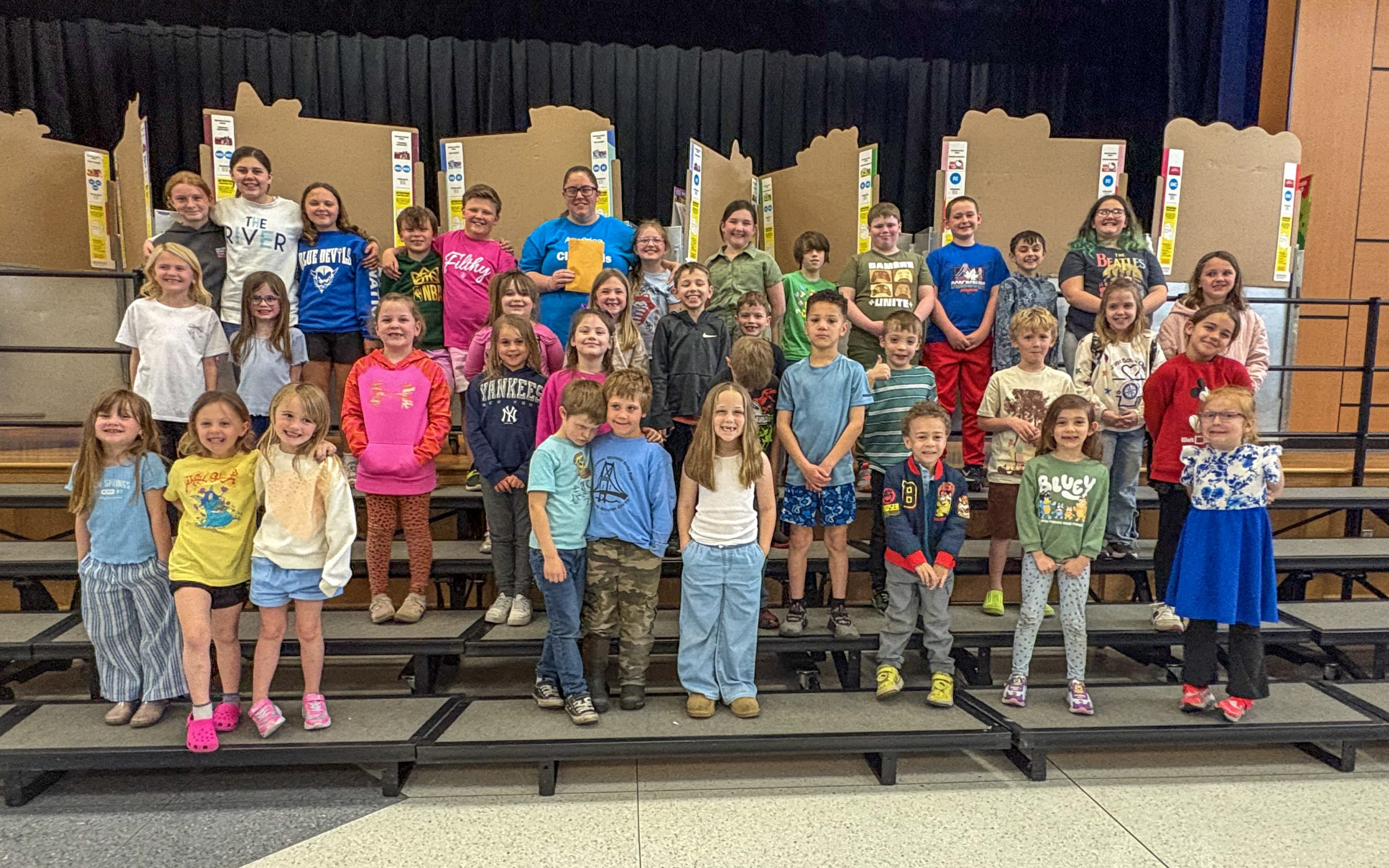Large group of young students standing on risers smiling with an adult female holding a brown envelope